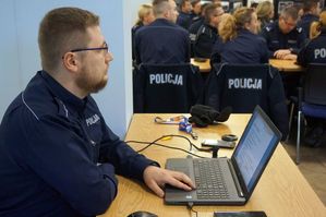 zdjęcie przedstawia umundurowanego policjanta, siedzącego przy laptopie, w tle natomiast są policjanci uczestniczący w szkoleniu