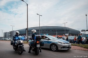 Zabezpieczenie meczu w Tychach. Policjanci na motocyklach służbowych oraz radiowóz oznakowany, w tle stadion