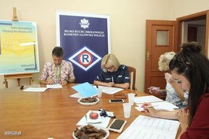 Komisja sprawdza prace konkursowe przy okrągłym stole. W tle znajduje się plakat Symbolu Ogólnopolskiego Policyjnego Dnia Odblasków 2019 oraz banner Biura Ruchu Drogowego Komendy Głównej Policji.