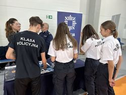 na zdjęciu młodzież stojąca przy stanowisku promocyjnym, rozmawiająca z umundurowanym policjantem z Wydziału Doboru i Szkolenia Komendy Wojewódzkiej Policji w Katowicach.