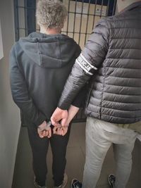 Na zdjęciu policjant pionu kryminalnego trzyma zatrzymanego mężczyznę, który ma założone kajdanki na ręce trzymane z tyłu.