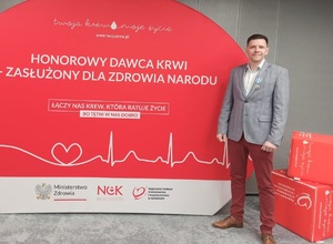 Na zdjęciu Łukasz Kaczyński stojący na tle planszy honorowy dawca krwi.