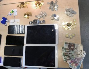 Na zdjęciu widać przedmioty skradzione przez włamywacza które leżą na stole, u góry są to kupki monet a na dole zdjęcia widać tablety, telefony komórkowe i pieniądze w gotówce