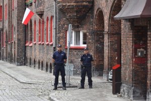 Na zdjęciu widać dwóch policjantów stojących przy bramie za nimi widać wiszącą flagę z okna