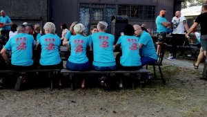 Fotografia kolorowa. Zdjęcie z pikniku charytatywnego. Osoby siedzą przy stole w koszulkach z logo rajdu motocyklowego.