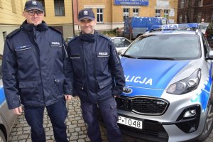Na fotografii widać dzielnicowych asp. szt. Ryszarda Prega oraz mł. asp. Tomasza Michalika, którzy stoją obok policyjnego radiowozu. W tle zdjęcia widać zaparkowane pojazdy.