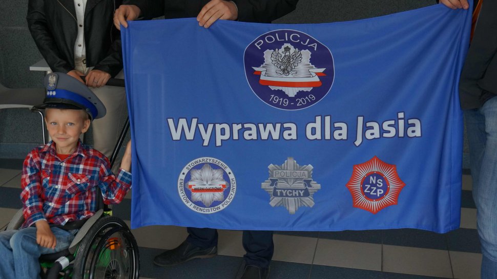 Na obrazku widoczny chłopczyk na wózku trzymający flagę z napisem WYPRAWA dla JASIA