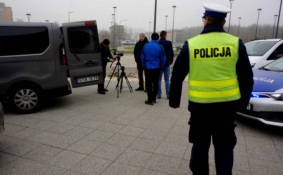 Na zdjęciu widoczny policjant ruchu drogowego z kamizelką odblaskową z napisem"policja" stoi tyłem, obok niego z prawej strony stoi zaparkowany radiowóz. Z lewej strony widoczy samochód z otwartym bagażnikiem obok 4 mężczyzn jeden zn ich ustawia kamerę na statyw. 