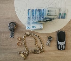 Zdjęcie kolorowe: na stole leży telefon komórkowy, zegarek, banknoty, złota biżuteria i klucze
