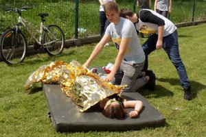 Zdjęcie z przebiegu testu udzielania pierwszej pomocy. Na materacu leży symulant obok niego zawodnik, który przykrywa go folią ochronną w kolorze złotym.